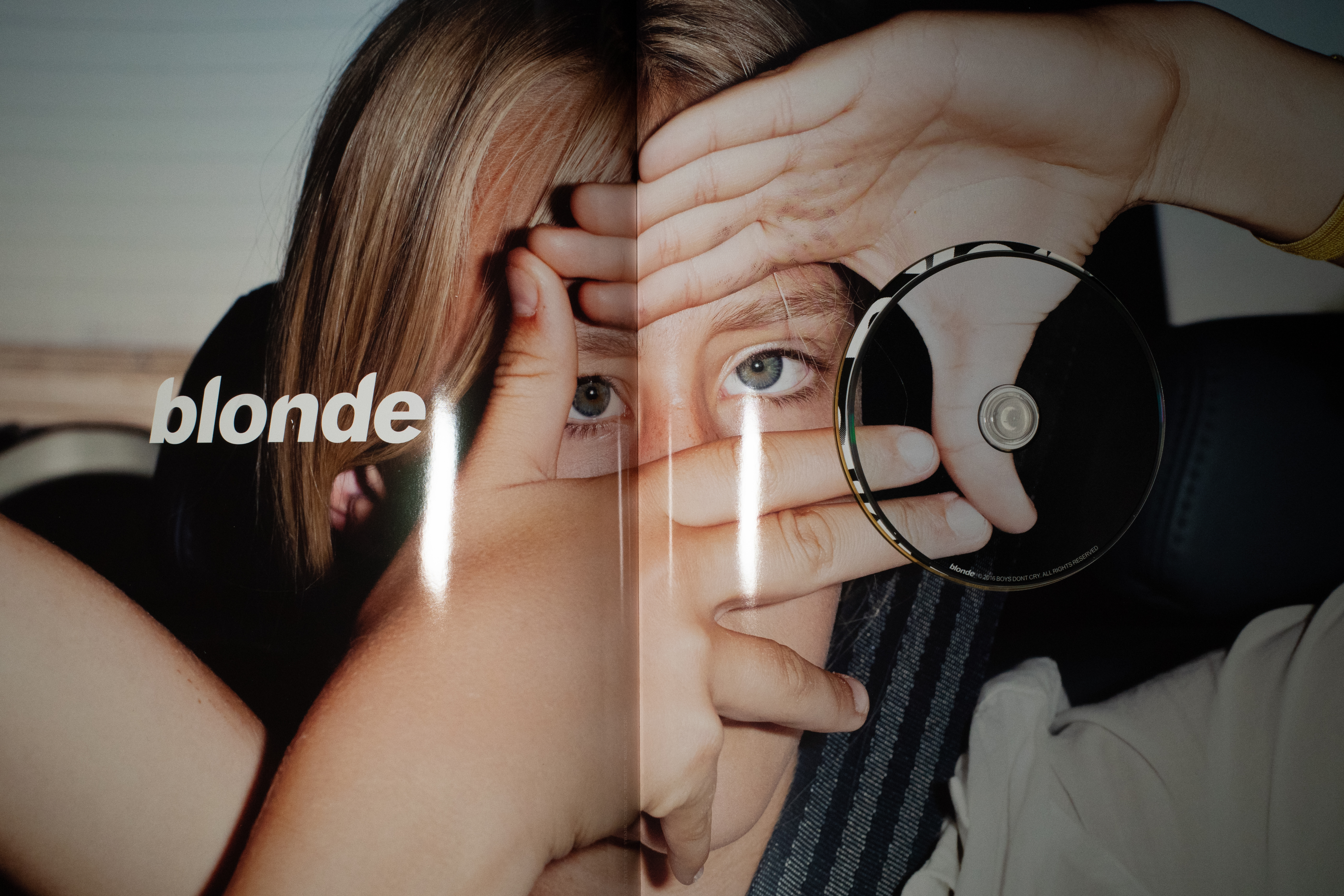 Image of a spread in the Boys Don't Cry Magazine. It features a blonde girl in the back of a car, with both her hands up to her face, framing her eyes with her fingers. On the left side of the spread, blonde is written in white italic text. On the right, there's a circle cutout with a Blonde album CD. The CD is printed with part of the image in the spread.