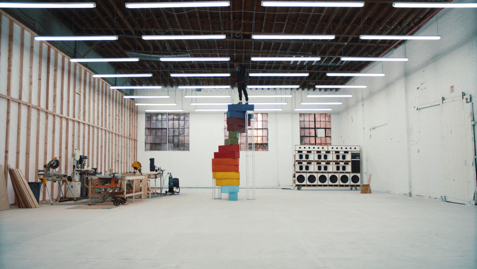 Frank Ocean standing atop a spiral staircase of colored steps in a large warehouse. On left is a workbench with powertools and wood. In the back are three large windows, and in the back right corner are many speakers. Overhead are many flourescent light tubes illuminating the whole space.